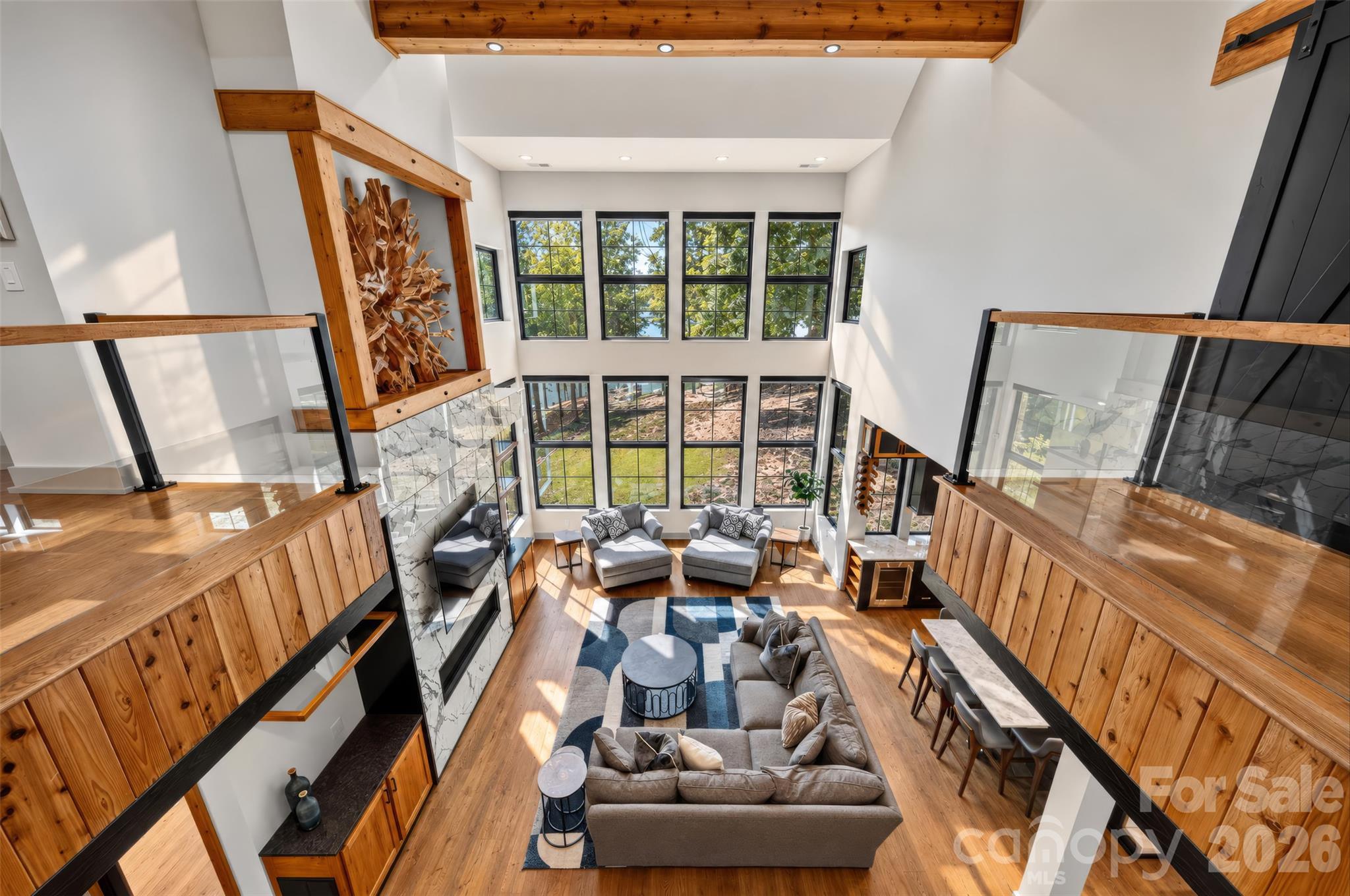 Double-height living room with tall windows, wood floors, and a sculptural wood wall feature.