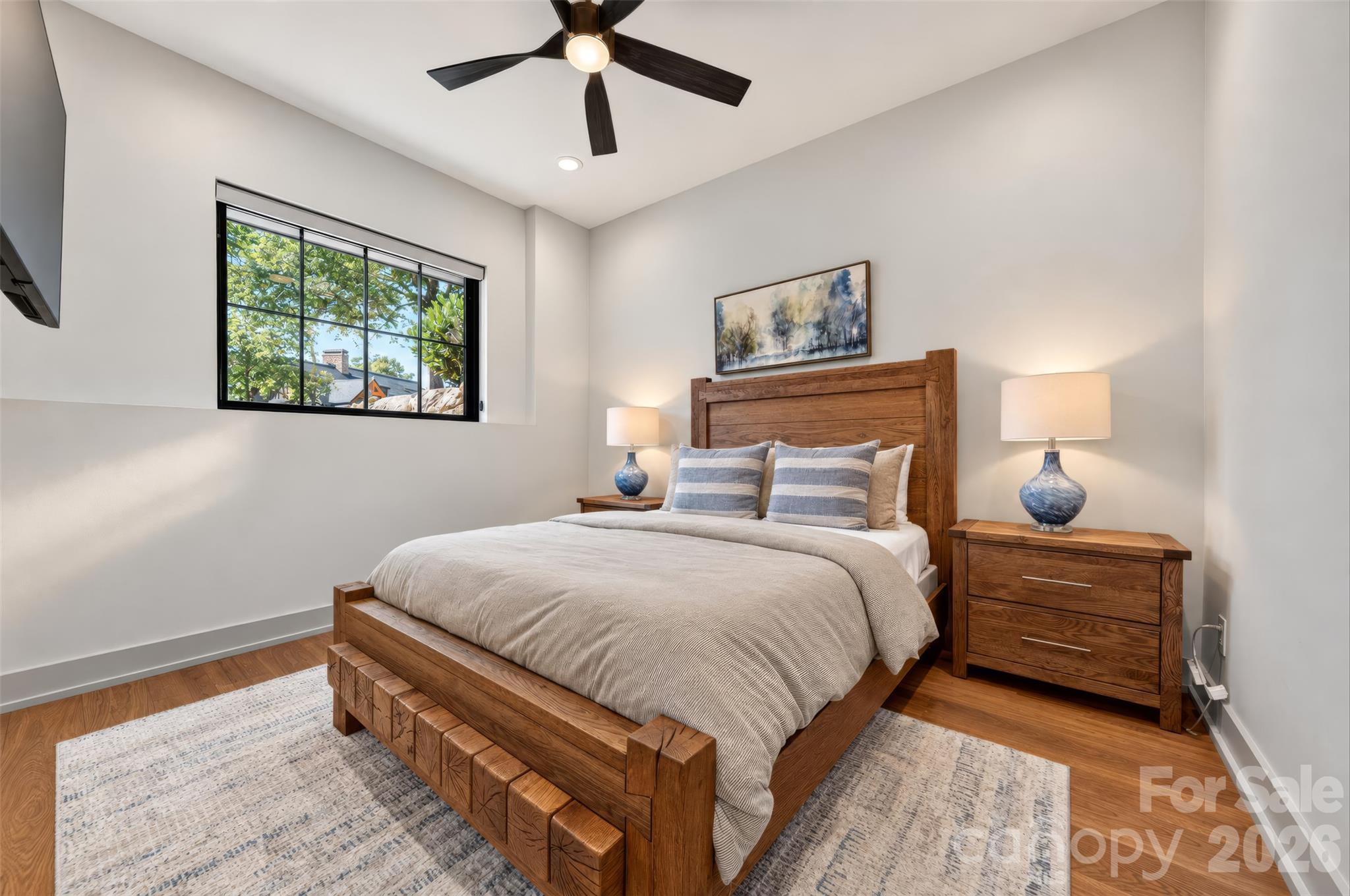 Bedroom with a large wooden bed, matching nightstands, and a window overlooking leafy trees.