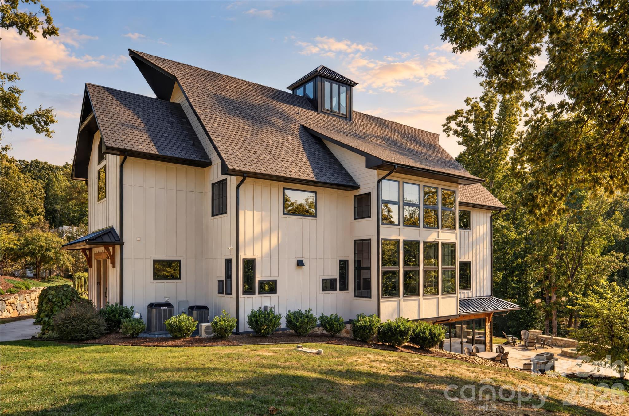 White modern farmhouse with a steep dark roof, tall windows, and a patio surrounded by trees.