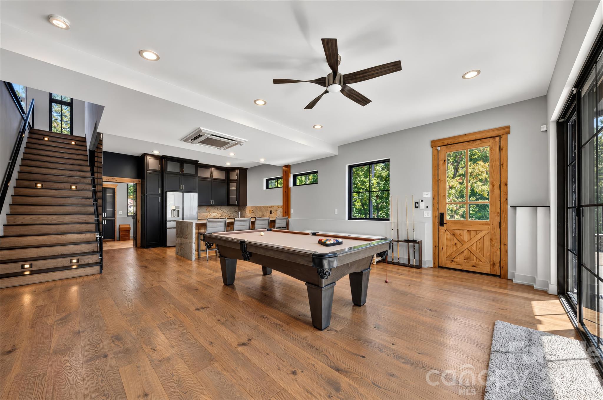 Game room with a pool table, wood staircase, dark kitchen cabinets, and large windows.