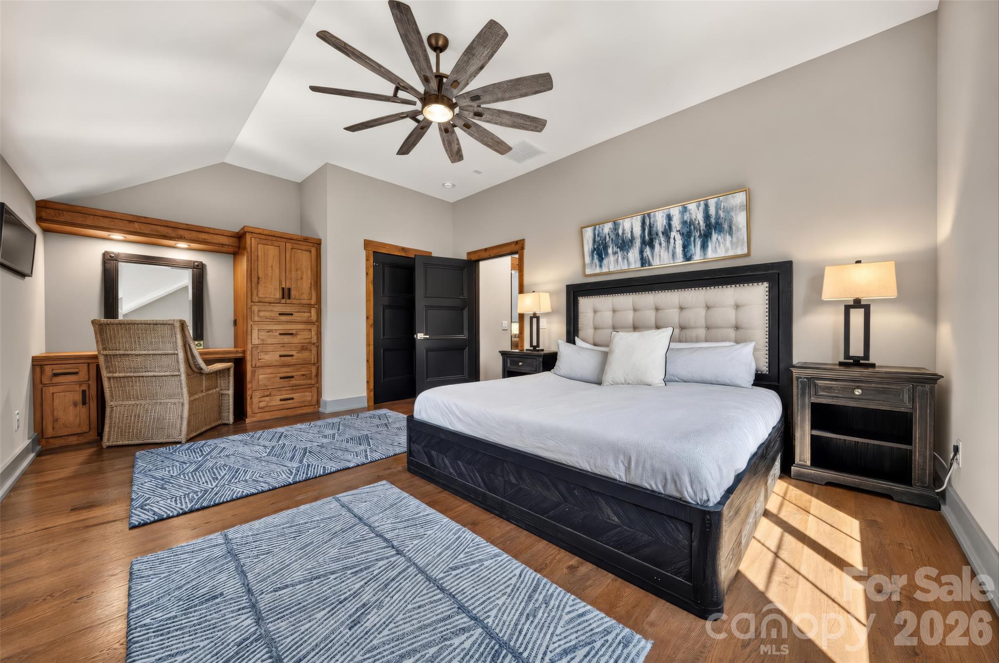 Furnished bedroom with a vaulted ceiling, dark-framed bed, wood cabinetry, and patterned blue-and-white rugs.