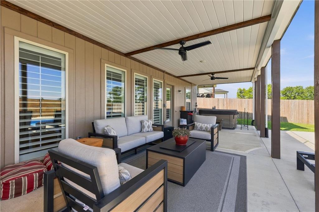 Covered patio with cushioned seating, ceiling fans, and a hot tub beside a fenced yard.