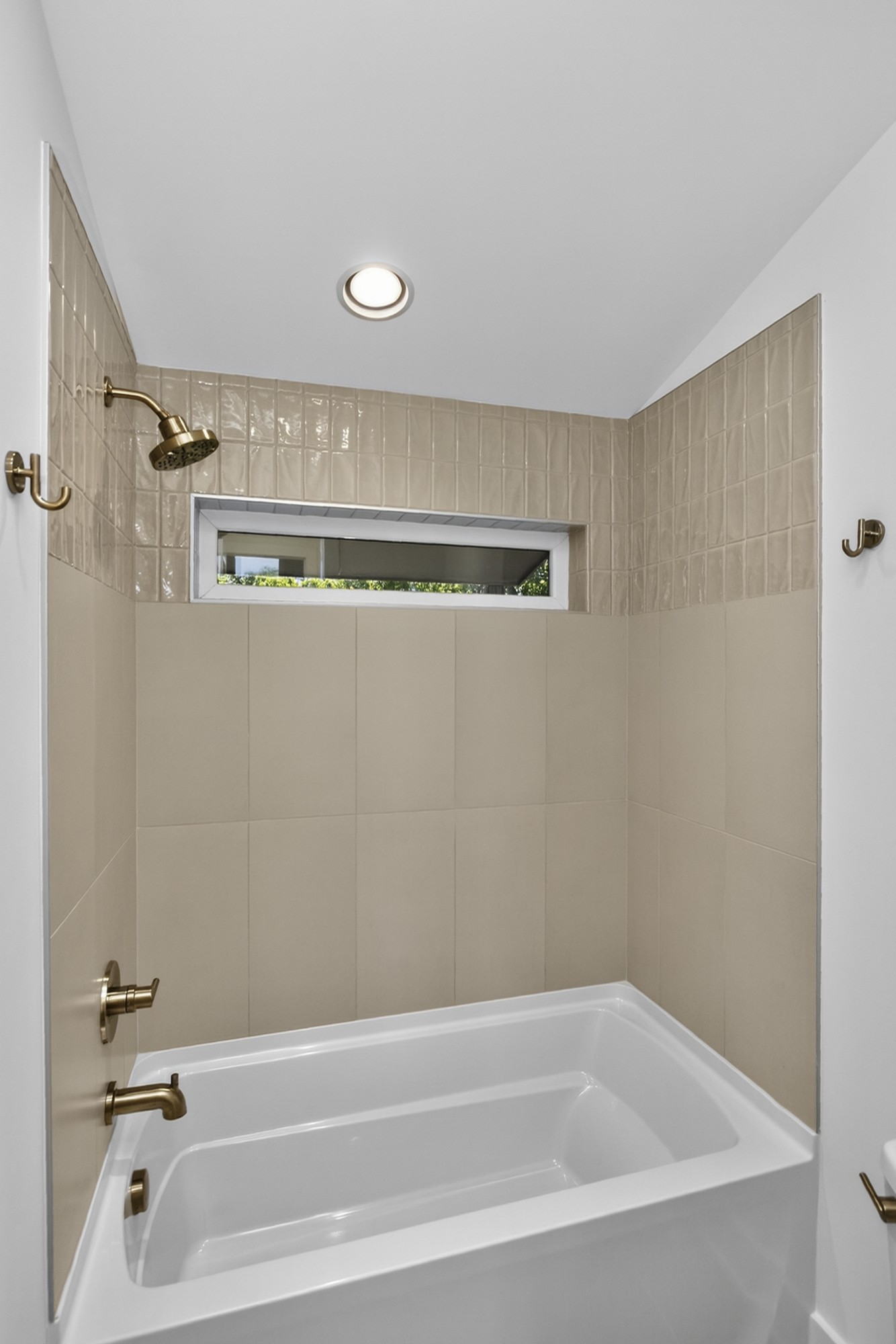 Bathroom tub and shower with beige tile, brass fixtures, and a narrow window.