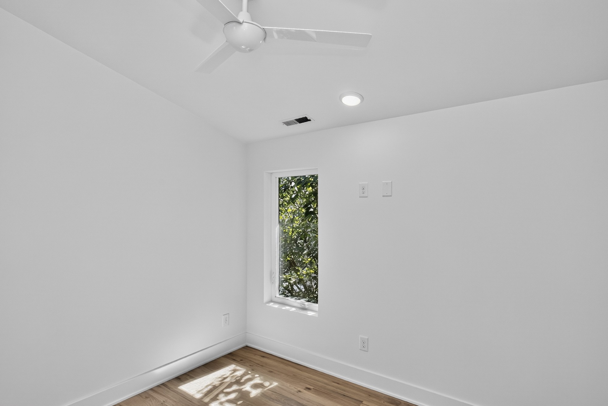 Vacant room with hardwood floors, a ceiling fan, and a narrow window overlooking greenery.