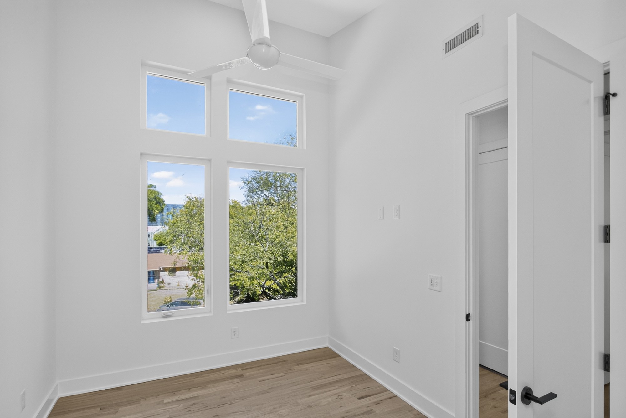 Vacant room with hardwood floors, a ceiling fan, and tall windows overlooking trees.