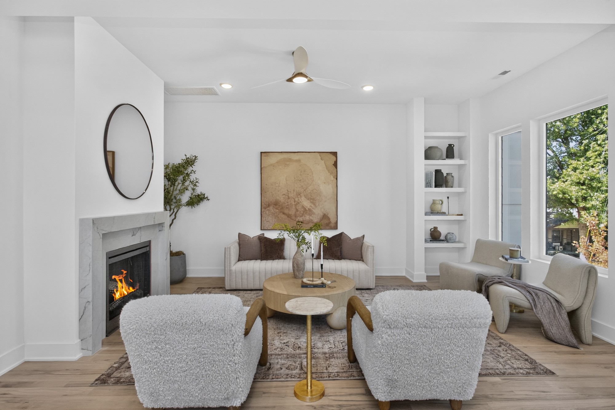 Living room with a marble fireplace, built-in shelves, and large windows overlooking trees.