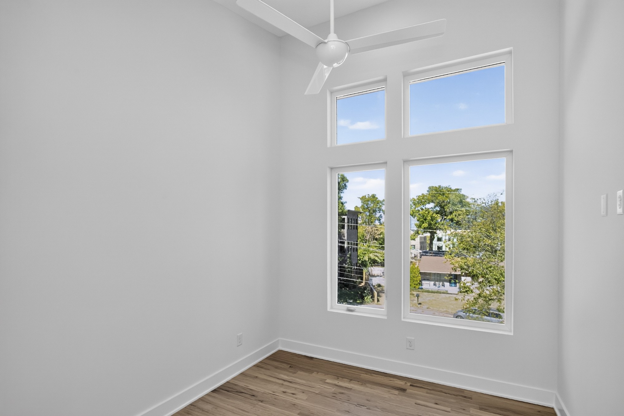 Empty room with hardwood floors, a ceiling fan, and tall windows overlooking leafy trees.