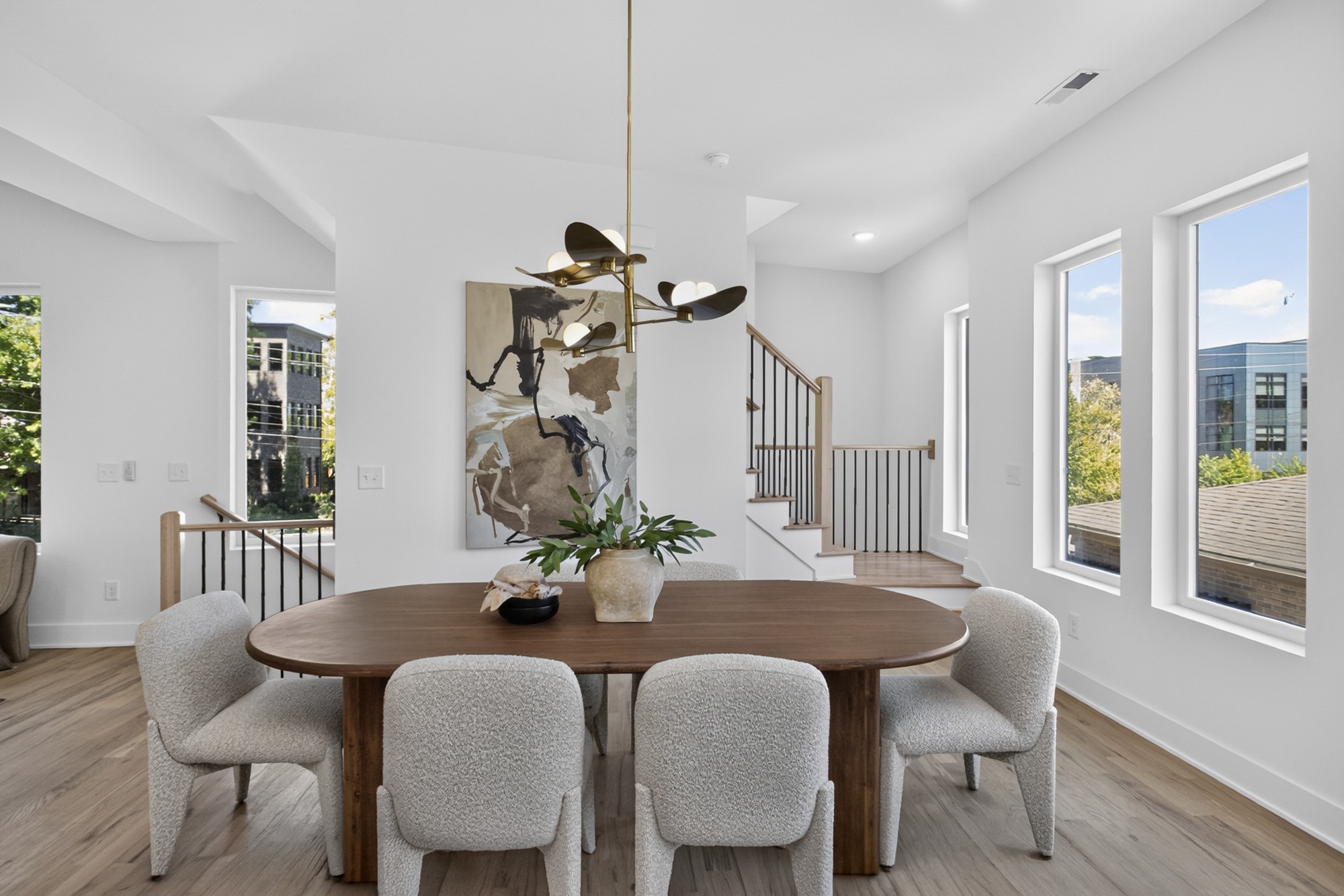 Dining area with a wood table, upholstered chairs, large windows, and an open staircase.