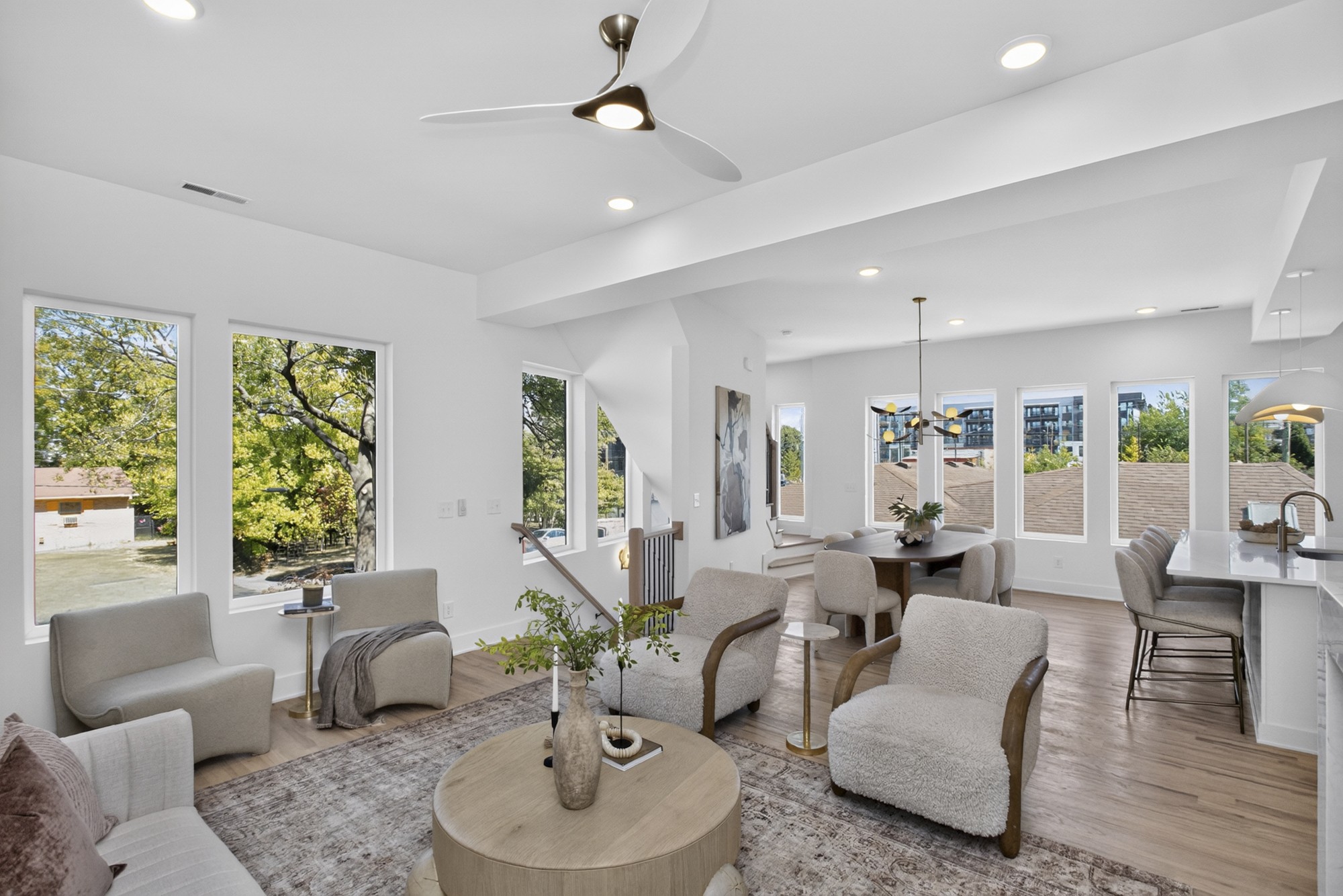 Open-plan living room with pale seating, large windows, wood floors, and an adjoining dining area and kitchen.