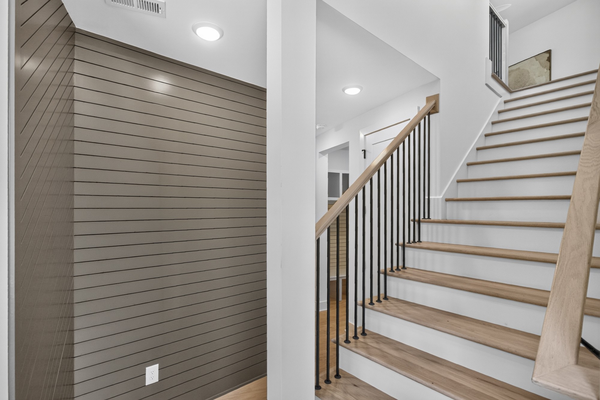 Modern staircase with light wood treads and black metal balusters beside a taupe paneled wall.