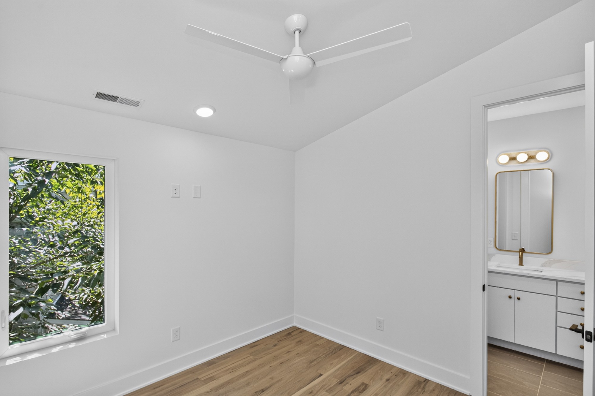 Empty bedroom with hardwood floors, a ceiling fan, leafy window view, and adjoining bathroom vanity.
