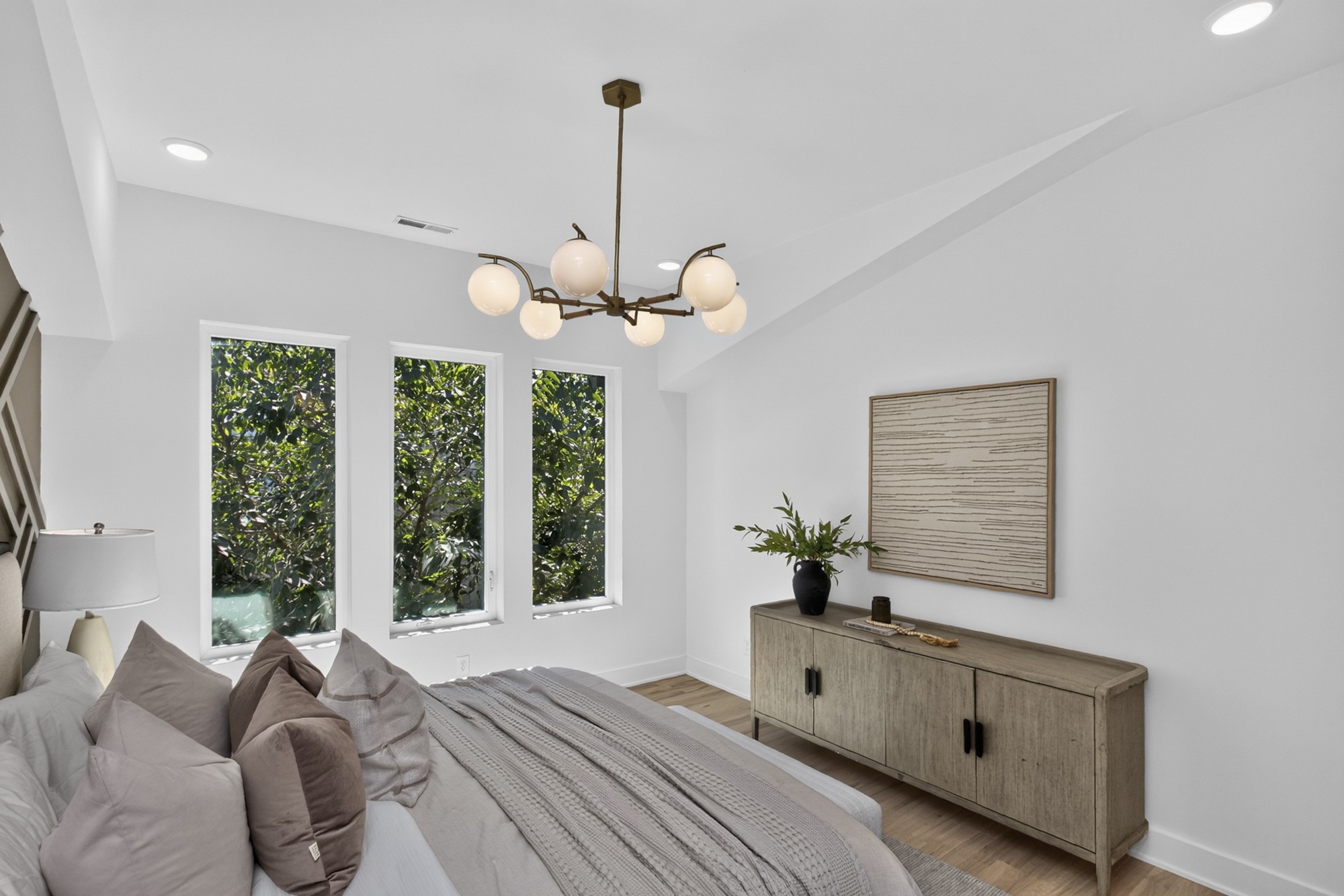 Bright bedroom with three tall windows overlooking leafy greenery and a wood sideboard beneath framed artwork.