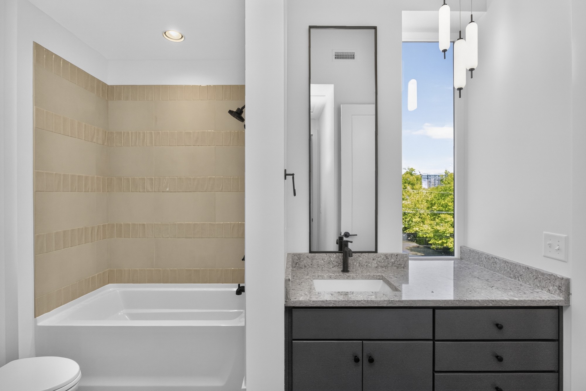 Bathroom with a beige tiled tub, gray vanity, stone countertop, and tall window.