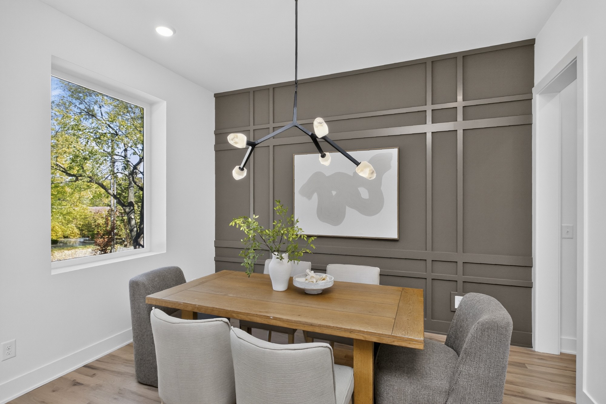Dining room with a wood table, paneled accent wall, and large window overlooking trees.