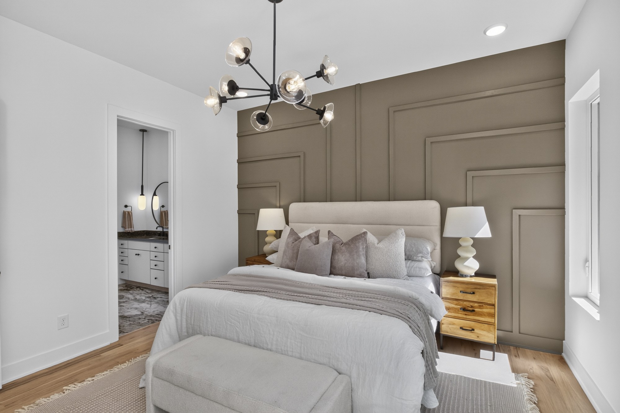 Staged bedroom with a geometric paneled accent wall, upholstered bed, and adjoining bathroom doorway.