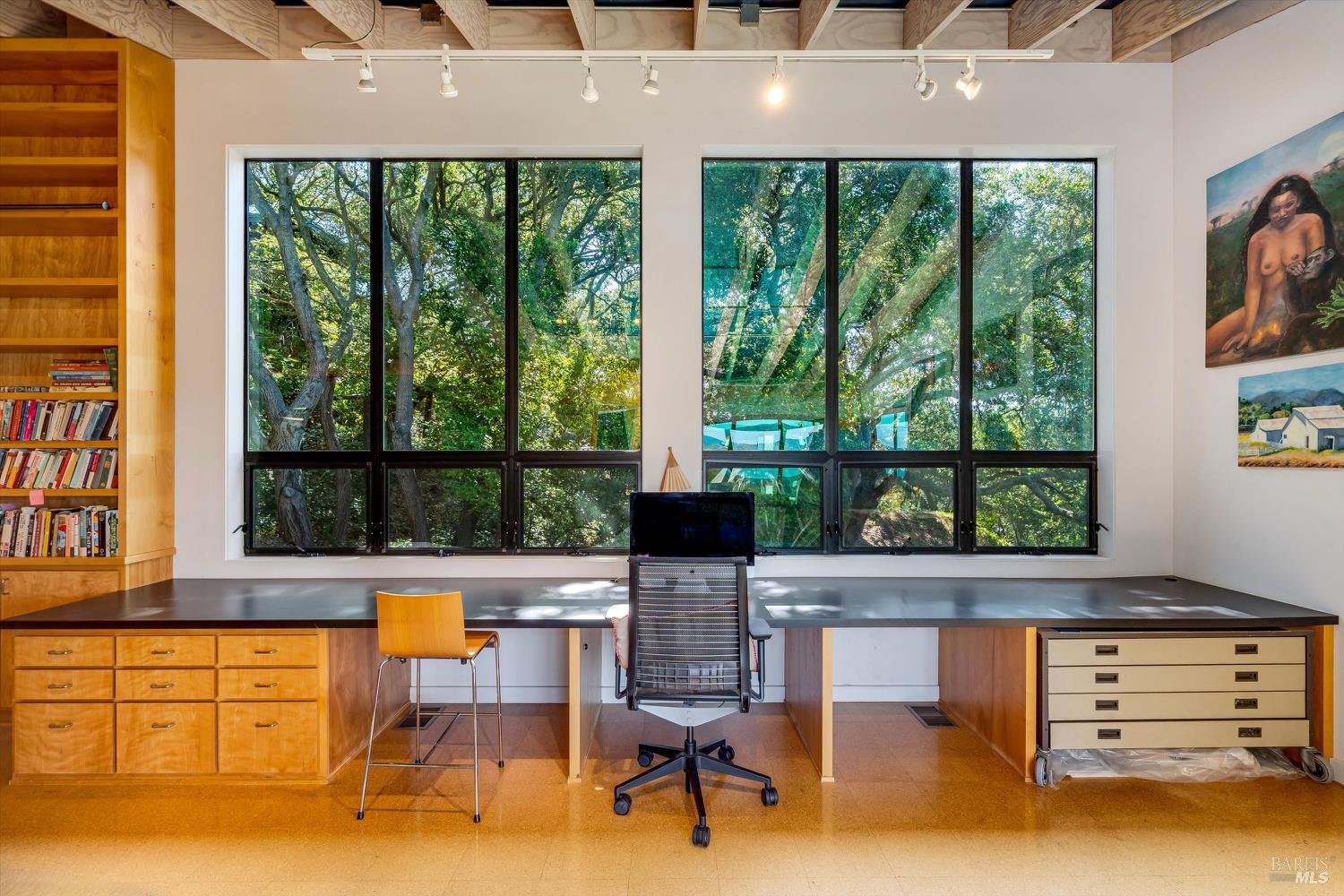 Home office with expansive windows overlooking trees, built-in desk, and exposed ceiling beams.