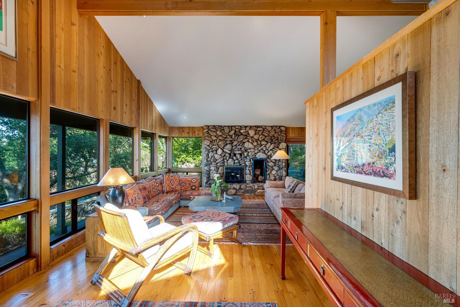 Living room with a stone fireplace, wood-paneled walls, and large windows overlooking greenery.