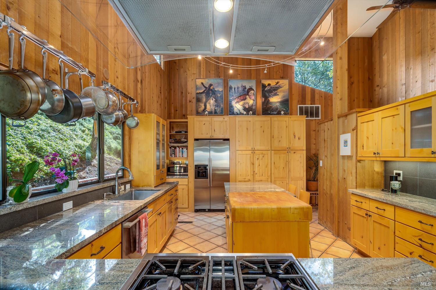 Wood-paneled kitchen with vaulted ceiling, central island, granite counters, and windows overlooking greenery.