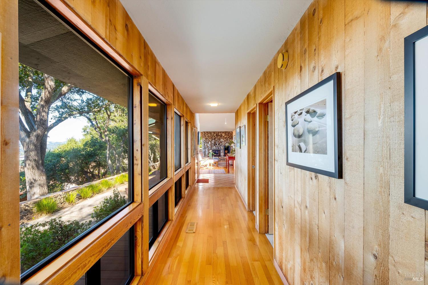 Wood-paneled hallway with hardwood floors, garden-facing windows, framed art, and a stone fireplace at the far end.