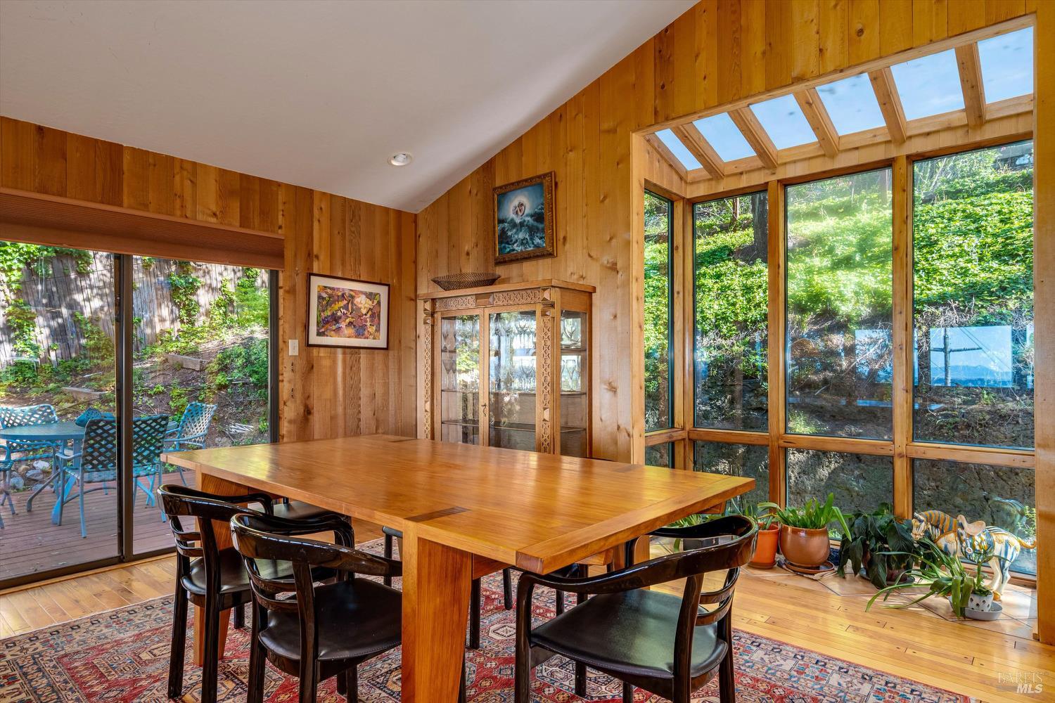 Dining room with a large wooden table, wood-paneled walls, and garden views through expansive windows.