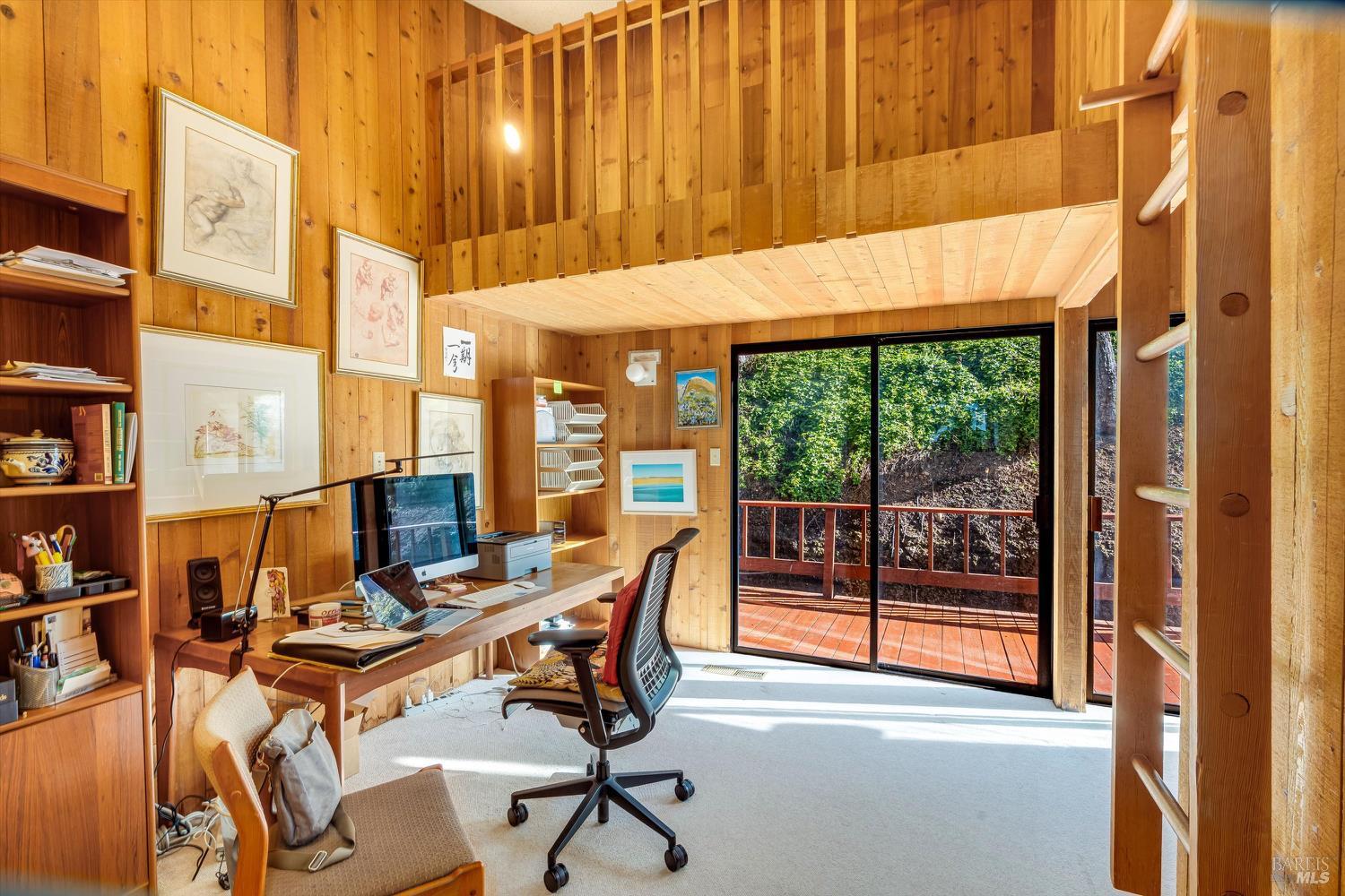 Wood-paneled home office with a desk, high ceiling, and sliding doors opening onto a deck.