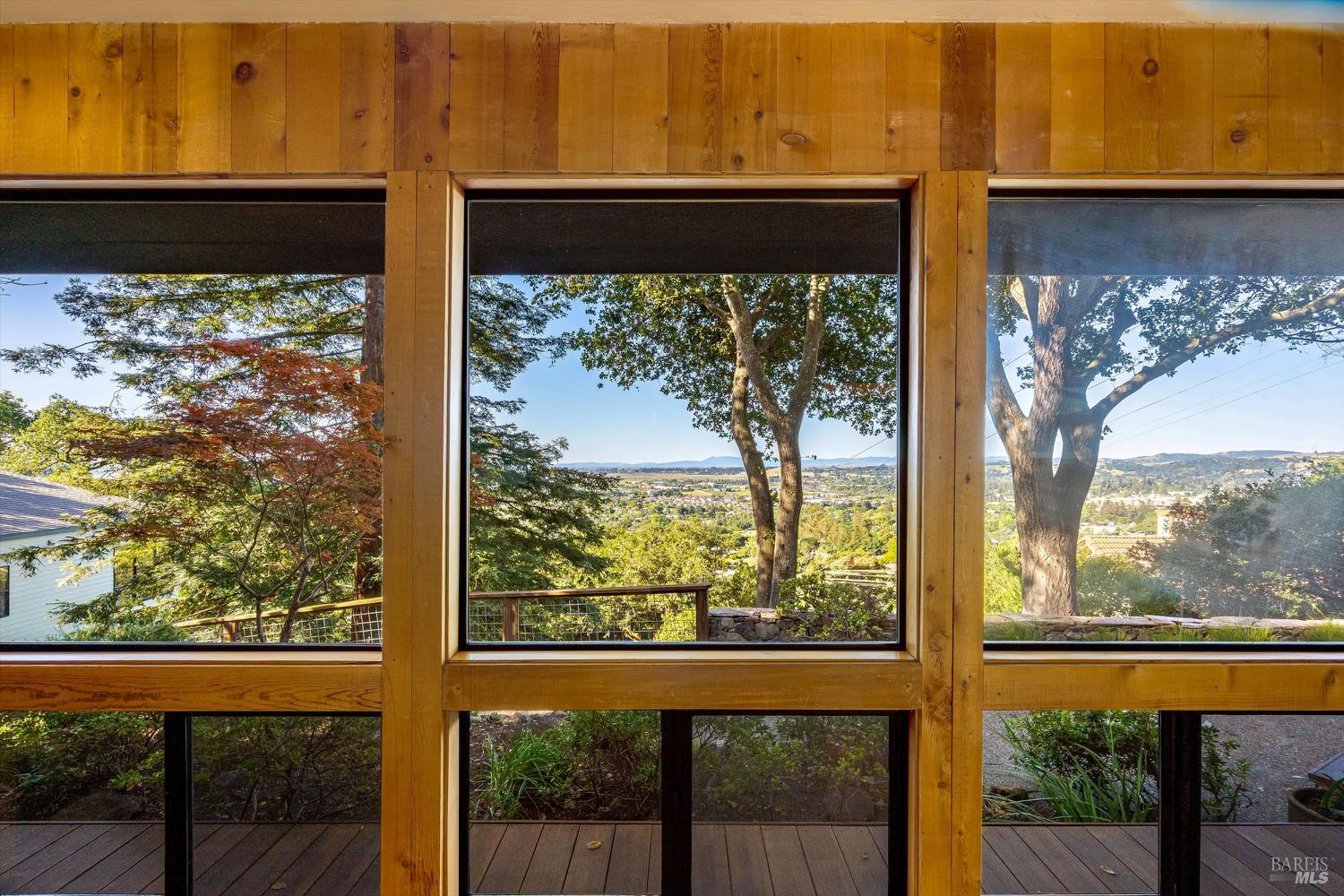 Valley view through large wood-framed windows, with mature trees and a deck outside.