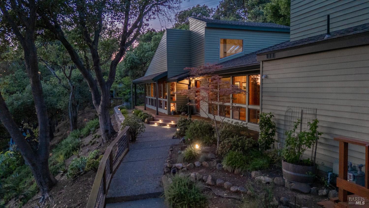 Multilevel gray home nestled among trees, with glowing windows and a lit garden path at dusk.