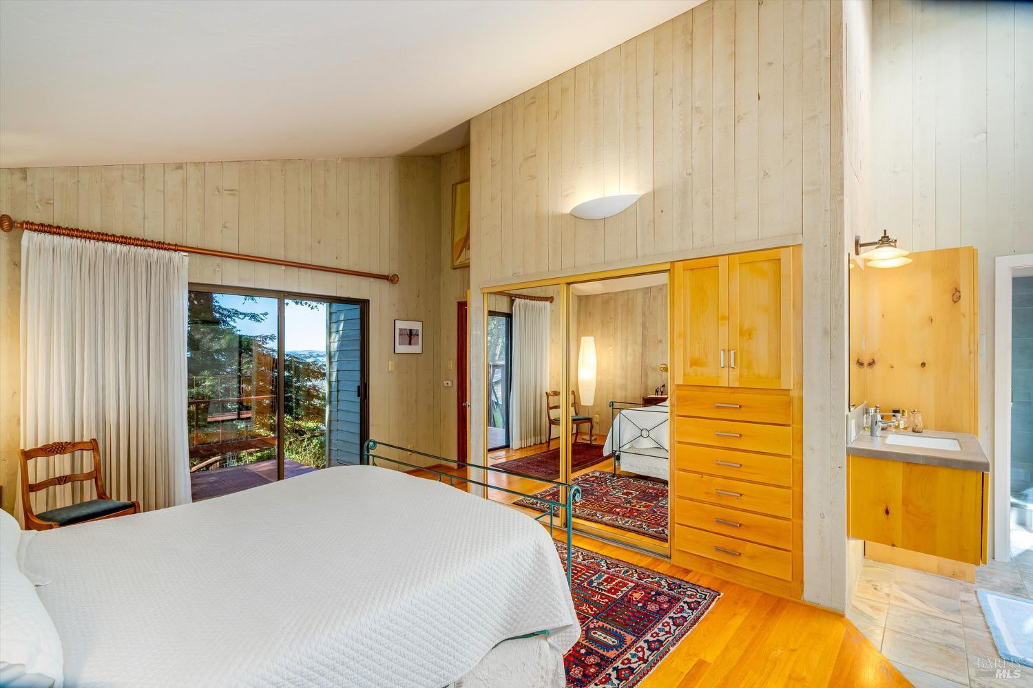 Bedroom with mirrored closet doors, wood built-ins, and a sliding glass door to a tree-lined deck.