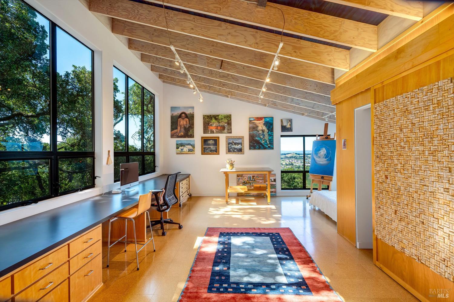 Bright home studio with vaulted wood ceiling, large windows, built-in desk, artwork, and easel.