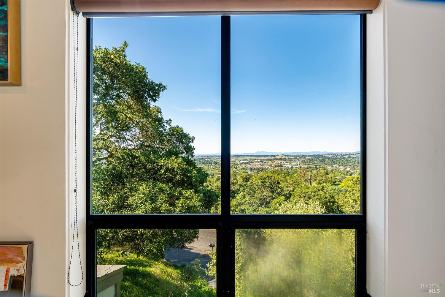 Valley and distant hills viewed through a large black-framed window, with leafy trees in the foreground.