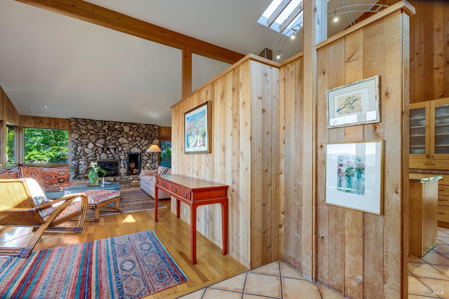 Living room with a stone fireplace, wood-paneled walls, patterned rugs, and a skylight.