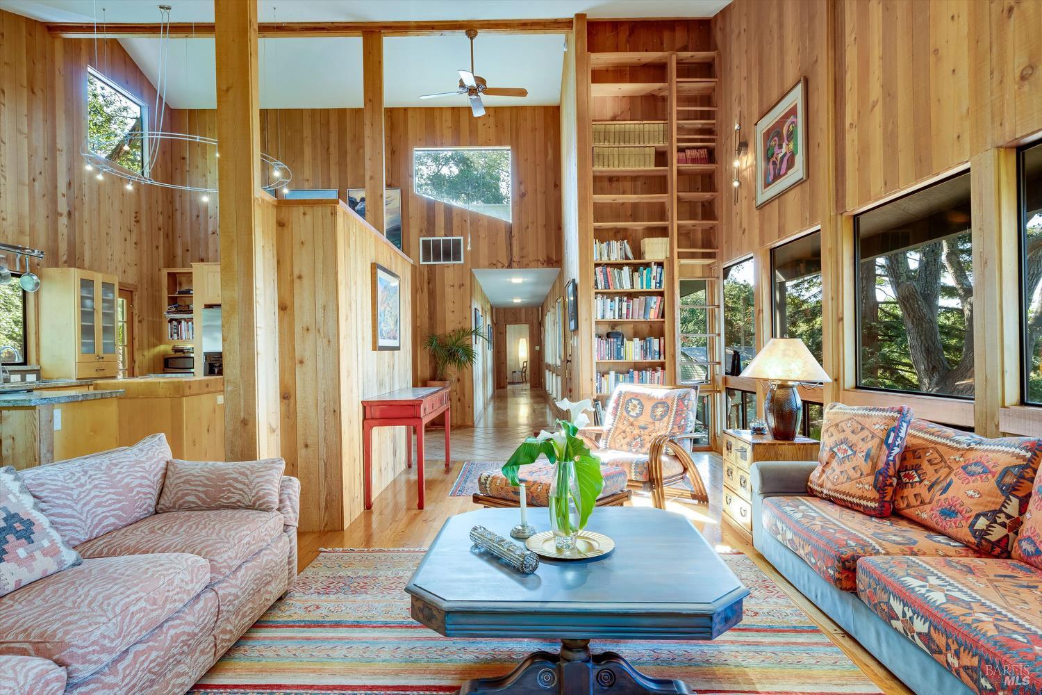 Wood-paneled living room with vaulted ceiling, tall bookshelves, and large windows overlooking trees.