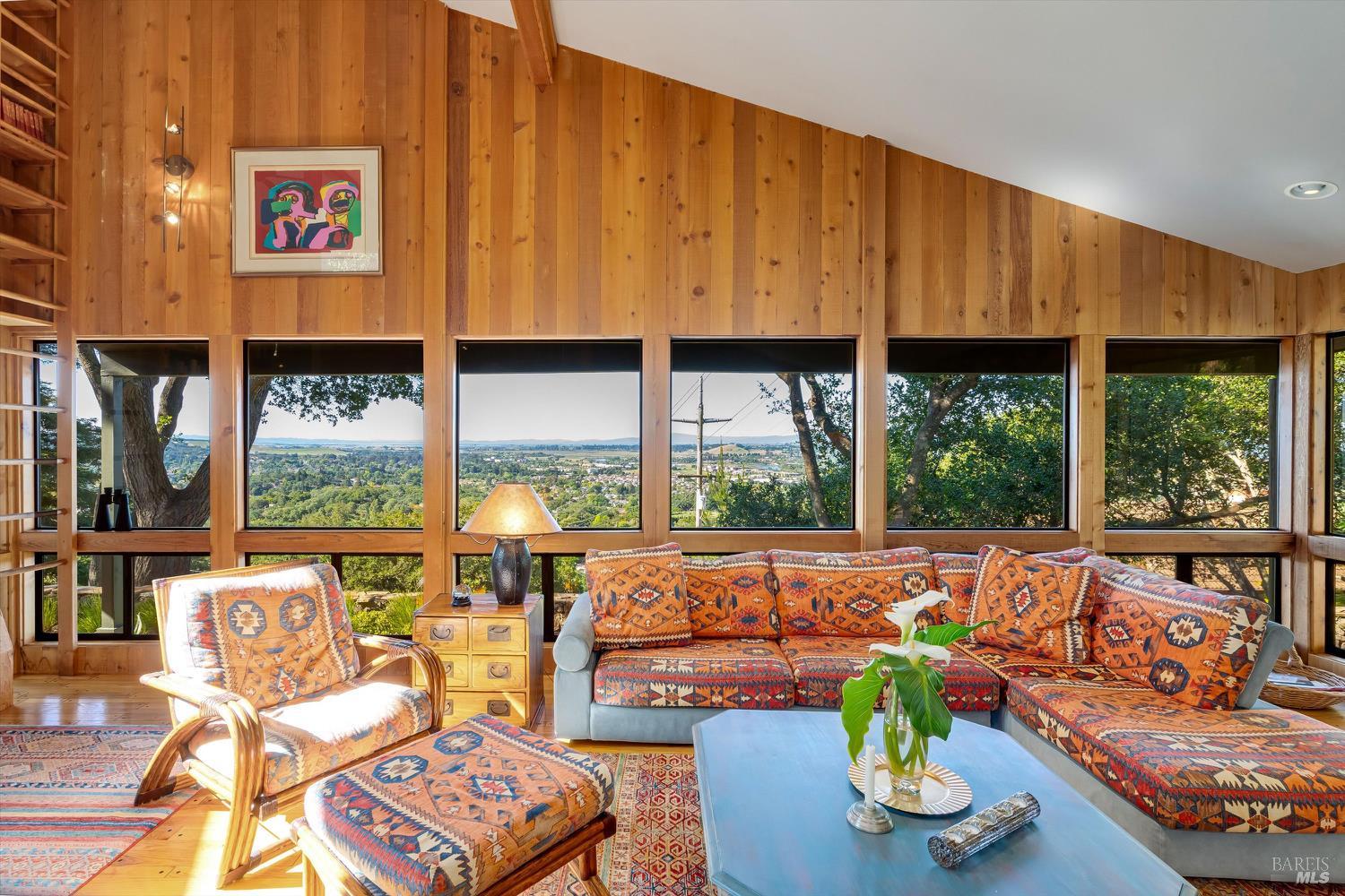 Wood-paneled living room with patterned seating and expansive windows overlooking a tree-filled valley.