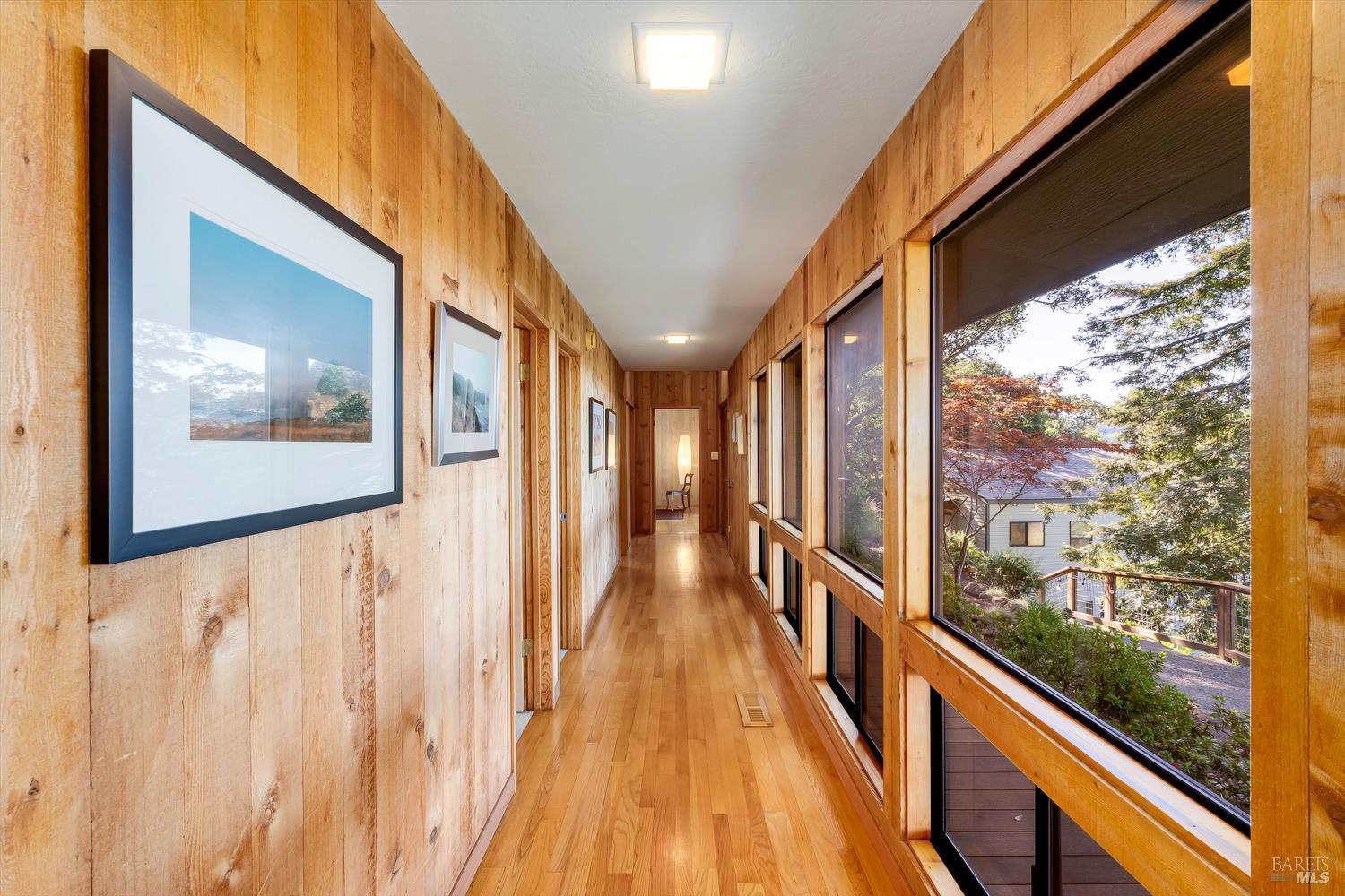 Wood-paneled hallway with hardwood floors, framed art, and large windows overlooking trees.