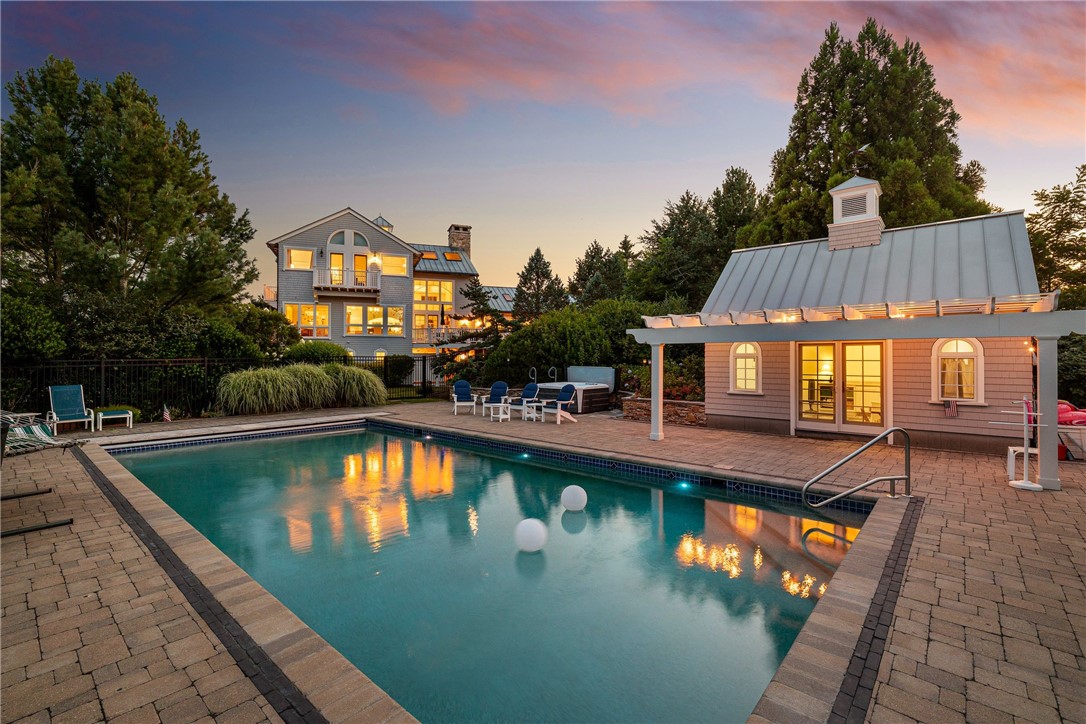 Outdoor pool and patio beside a lit pool house, with the home glowing among mature trees at dusk.