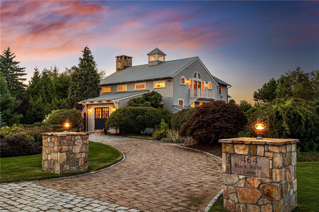 Gray home with a cupola behind a curved stone driveway and entrance pillars at dusk.