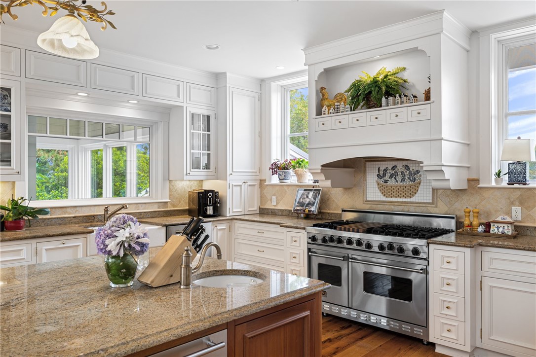 Bright white kitchen with granite island, stainless steel range, and windows overlooking greenery.