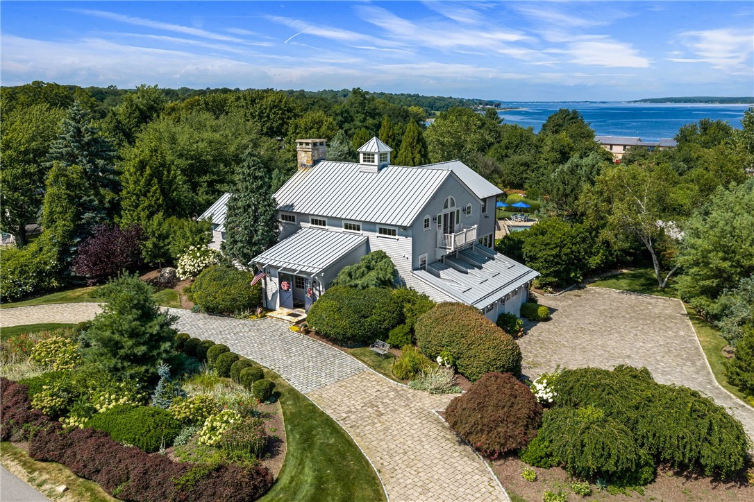 Aerial view of a gray coastal home with a metal roof, landscaped grounds, and water beyond the trees.