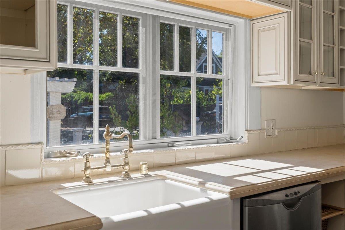 Kitchen sink beneath a large sunlit window, with a brass faucet and pale tiled backsplash.