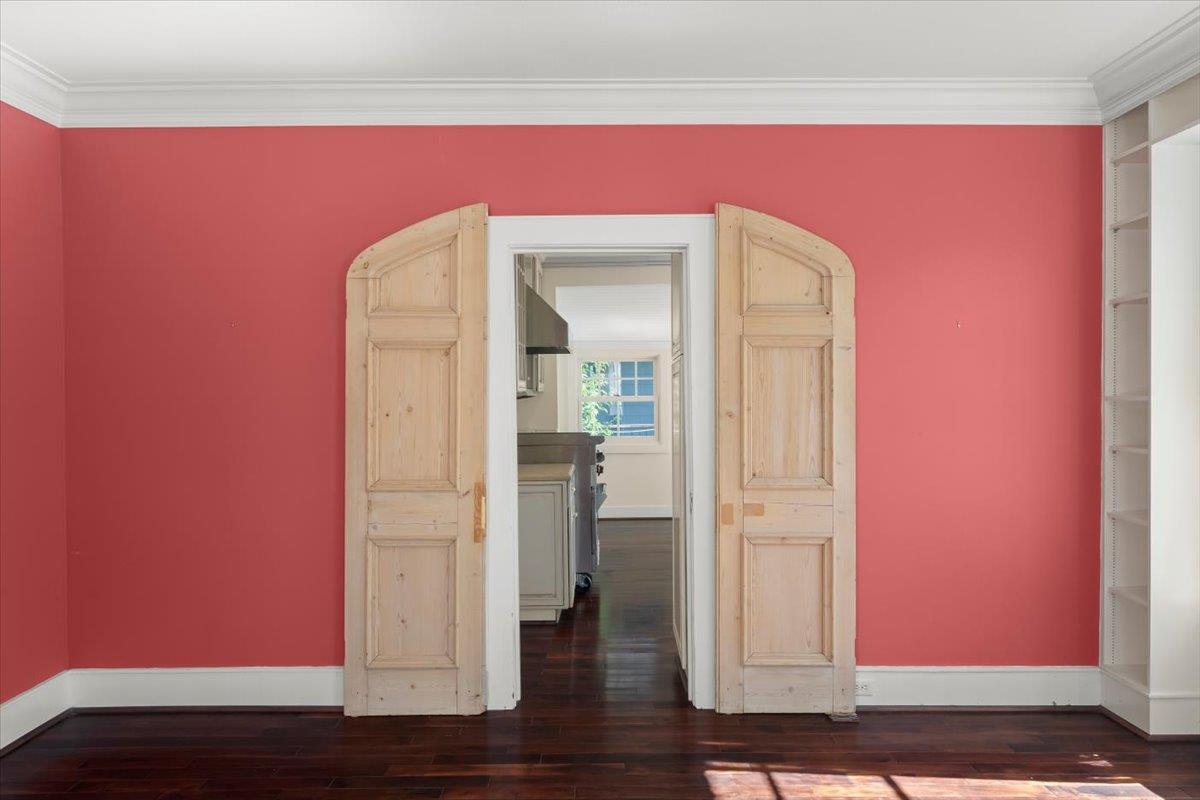 Empty coral-red room with dark hardwood floors and open wooden double doors leading to a kitchen.