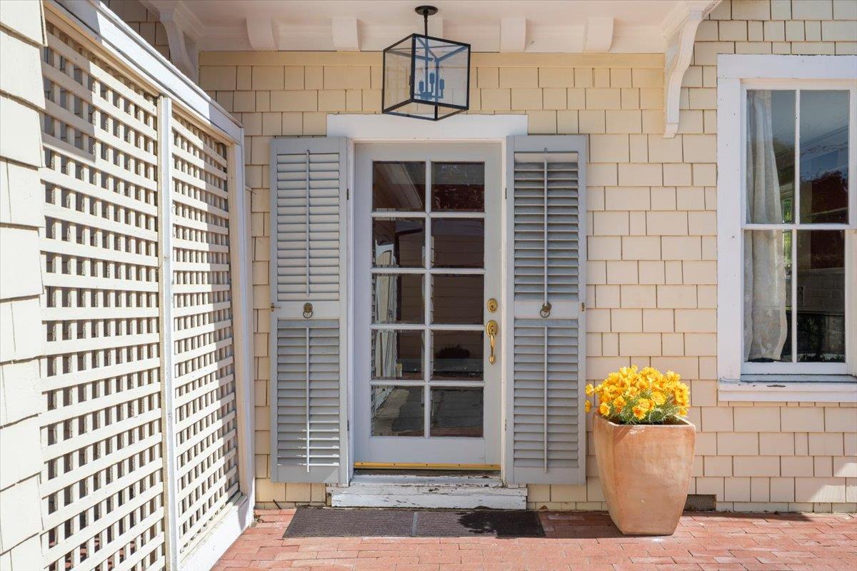 Front entrance with a glass-paneled door, white shutters, lattice screen, and potted yellow flowers.