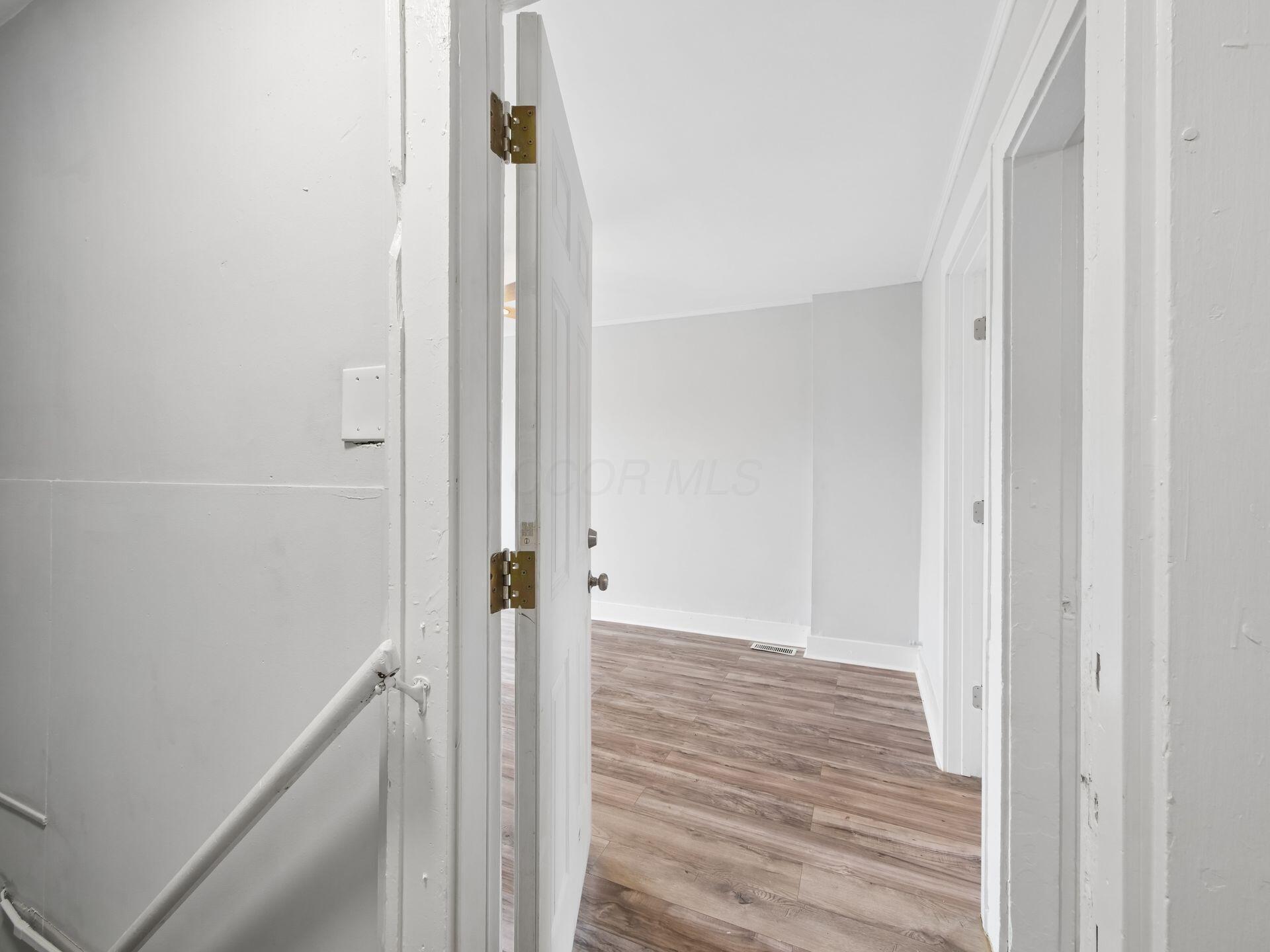 Empty room with wood-look flooring viewed through an open doorway, with a stair railing in the foreground.