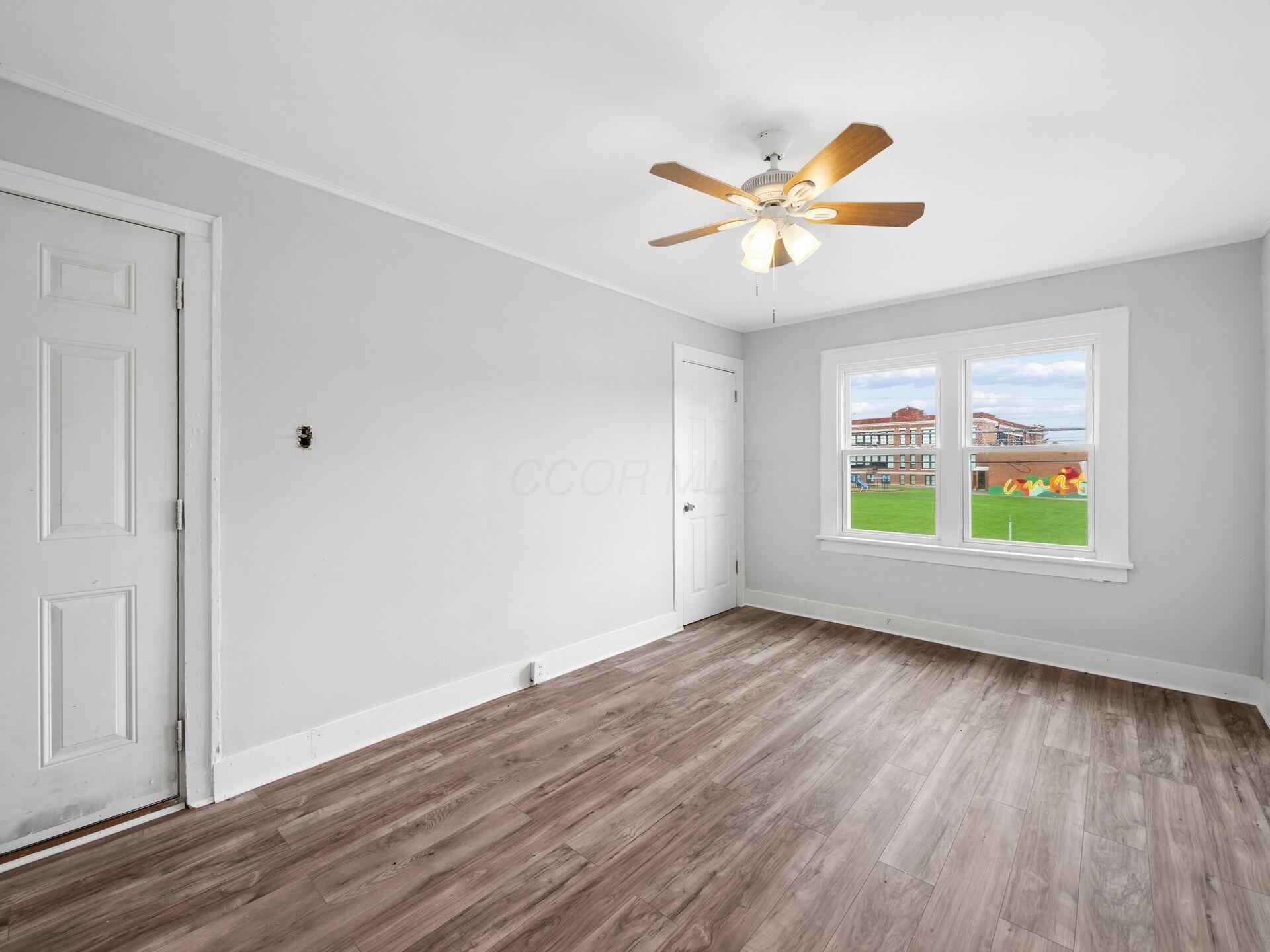 Empty bedroom with wood-look flooring, a lit ceiling fan, and two windows overlooking a lawn.
