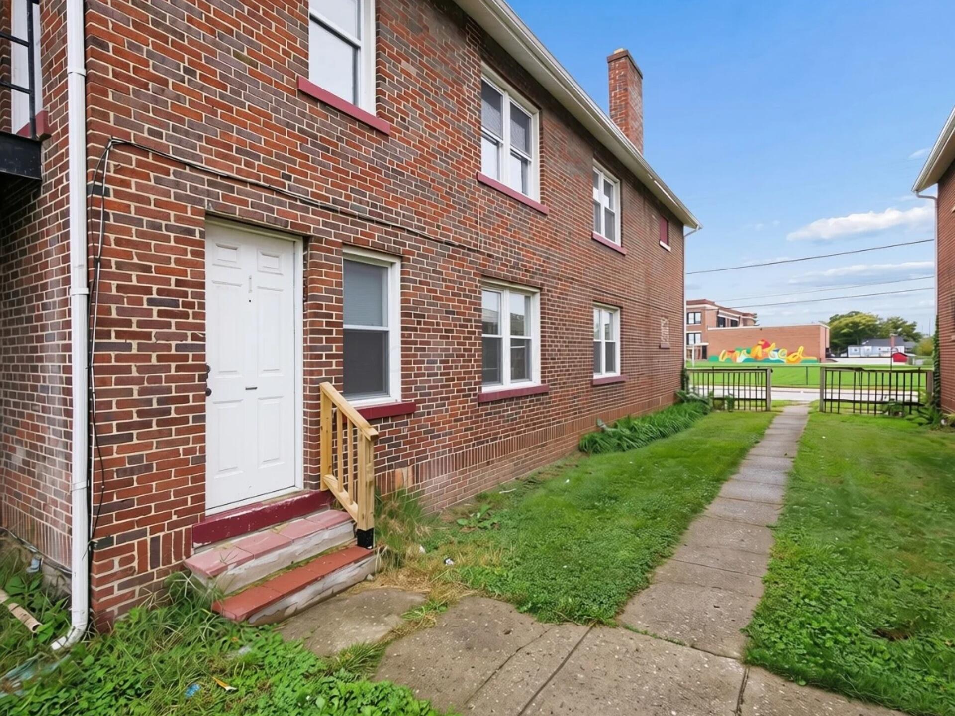 Two-story brick building with a white entry door, short steps, and a walkway through the grass.