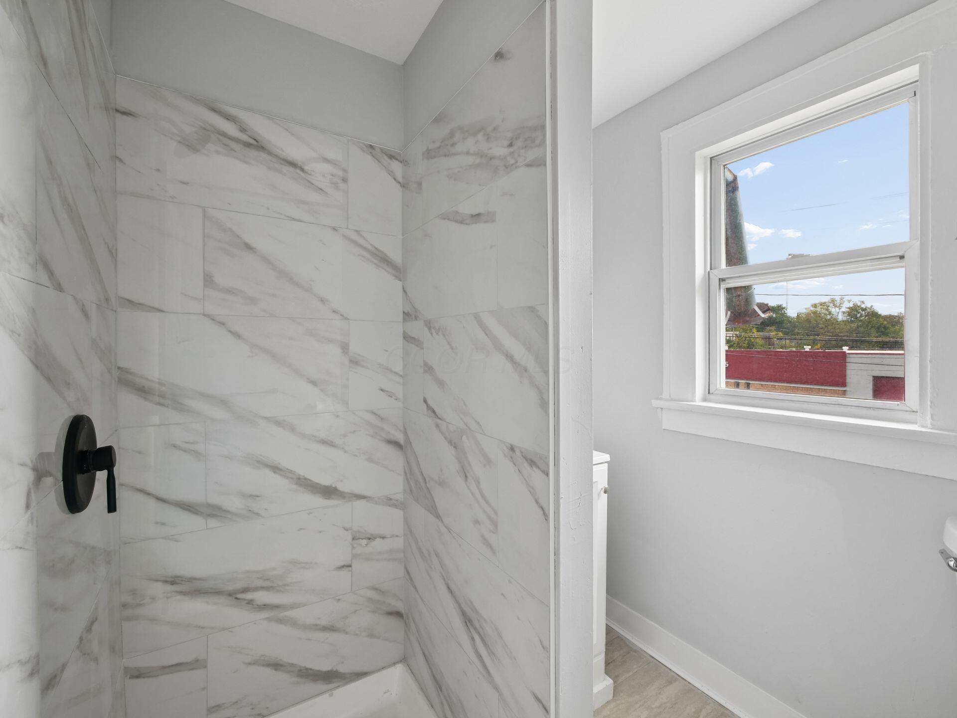 Bathroom with a marble-look tiled shower, black fixture, and window overlooking nearby rooftops.