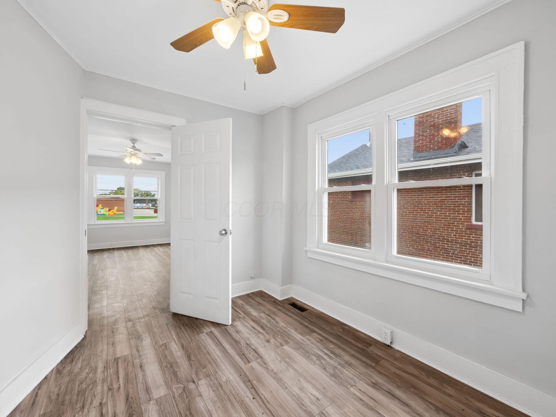 Empty room with wood-look flooring, a ceiling fan, and large windows beside an open doorway.