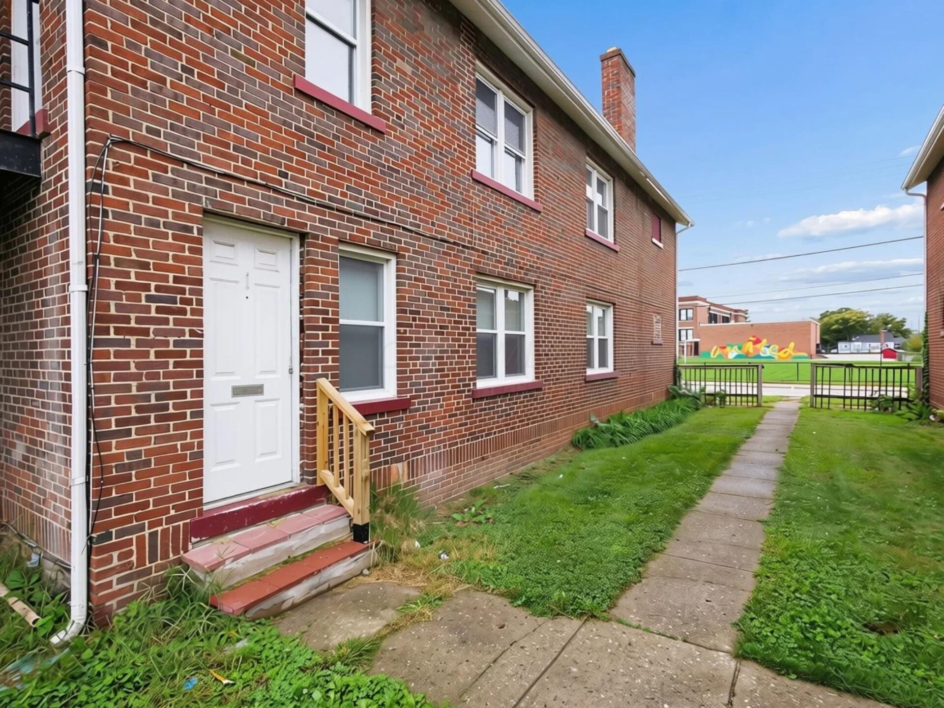 Red-brick building exterior with a white entry door, wooden railing, and walkway through a grassy side yard.