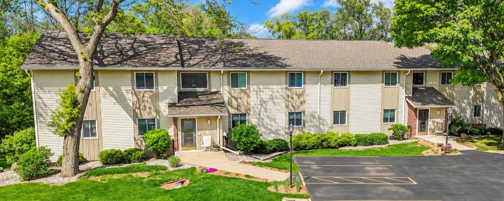 Two-story beige residential building with multiple entrances, leafy trees, and a parking lot.