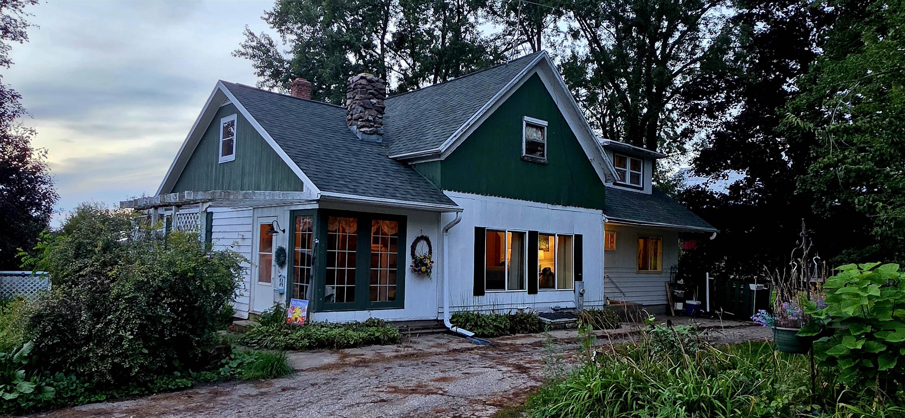 Green-and-white gabled house with a stone chimney, gravel drive, and lush garden beds.