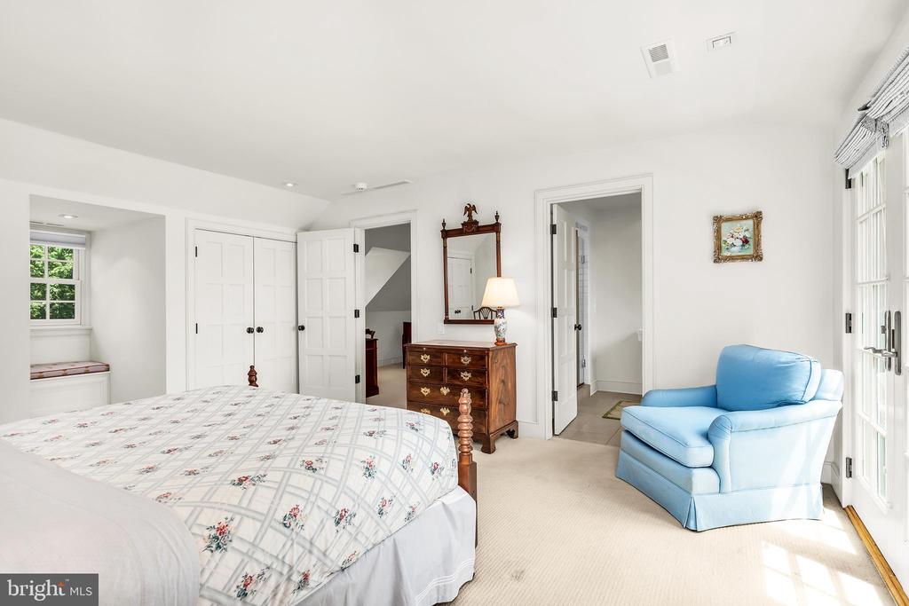 Furnished bedroom with a floral bed, blue armchair, wooden dresser, and bright windows.